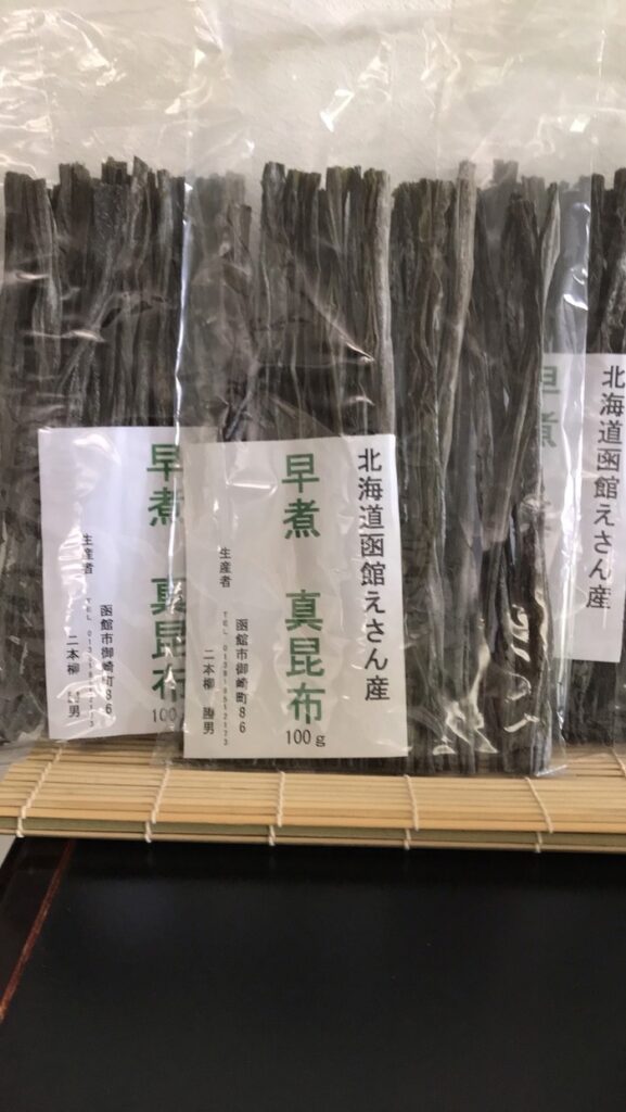 「早煮昆布」と書かれた100g入りの乾燥昆布のパッケージが、棚に縦にきれいに並べられている。パッケージには日本語で書かれたラベルが付いており、北海道恵山の品質の高さをアピールしている。.