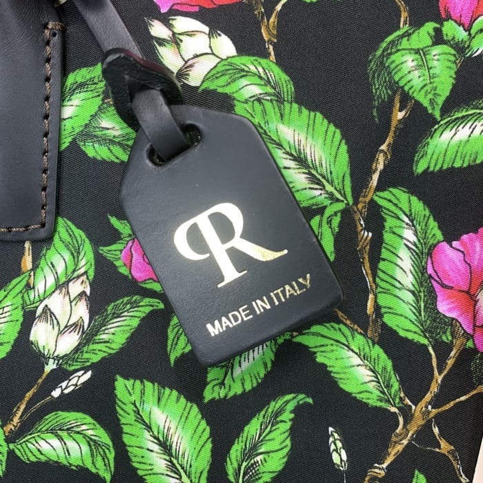 Eticheta neagră de bagaj ROBERTA PIERI cu "R" auriu și text italian este suspendată de o țesătură florală cu frunze verzi și flori roz, completând mini duffel cu eleganță italiană atemporală.