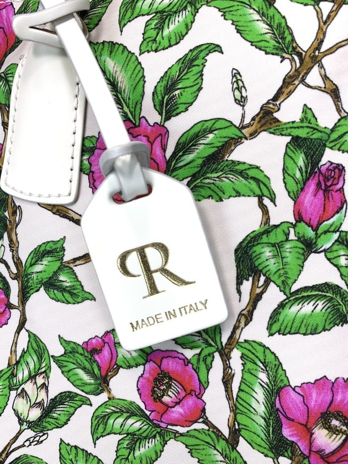 Prim-plan al geantei ROBERTA PIERI. pedunculi cu frunze verzi și flori roz; Eticheta albă atașată la geanta cu 2 căi are un logo auriu "R" și cuvintele "MADE IN ITALY".